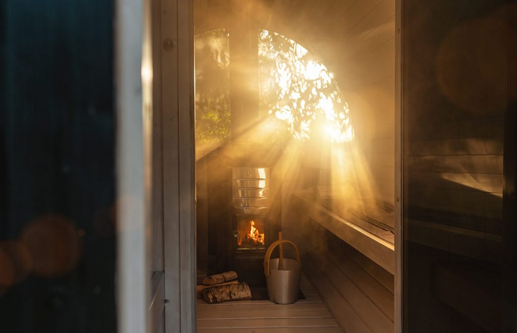 Ein Bild von Seppo Sauna zeigt ein Fasssauna von SEPPO SAUNA im Sonnenuntergang mit einer Aufgusszeremonie. Durch das große Panoramafenster bietet sich eine wunderschöne grüne Aussicht. Die Sauna wird mit Feuer beheizt, und auf dem Boden sind ein Eimer und eine Kelle zu sehen.